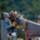A construction worker welding a steel beam outdoors, emitting sparks, wearing safety gear.